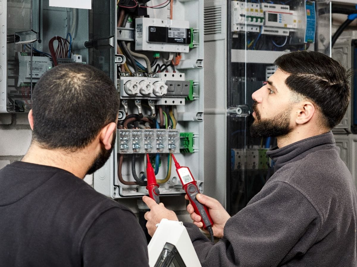 Zwei Elektriker von Miton messen an einer geöffneten Zählerverteilung; einer hält den Zweipol-Prüfer an die Klemmen.